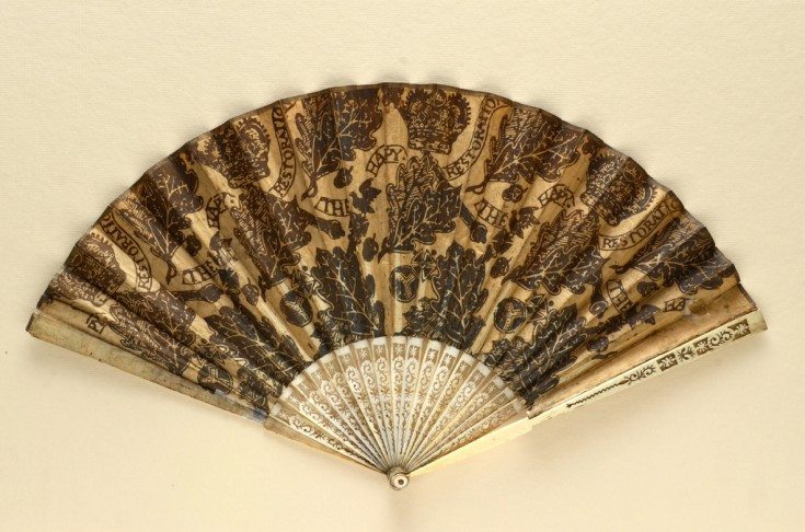 Folding fan made to commemorate the restoration of King Charles II to the throne of England. The paper mount is adorned on both sides with woodcut prints of oak leaves, acorns, orbs, sceptres and crowns; scrolls bearing the inscription ‘The Hapy Restoration’ weave in and out of the main design. English, c.1660 The Fan Museum, HA1723 